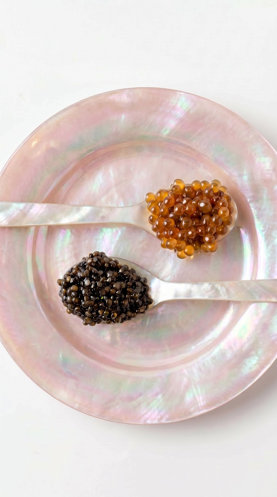 Two mother-of-pearl spoons of caviar, one dark and one amber, on a blush pearl plate