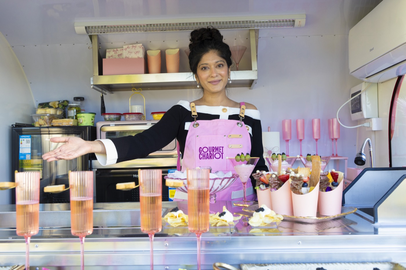 Nisha Singh hosting from the Chariot window in a pink Gourmet Chariot apron